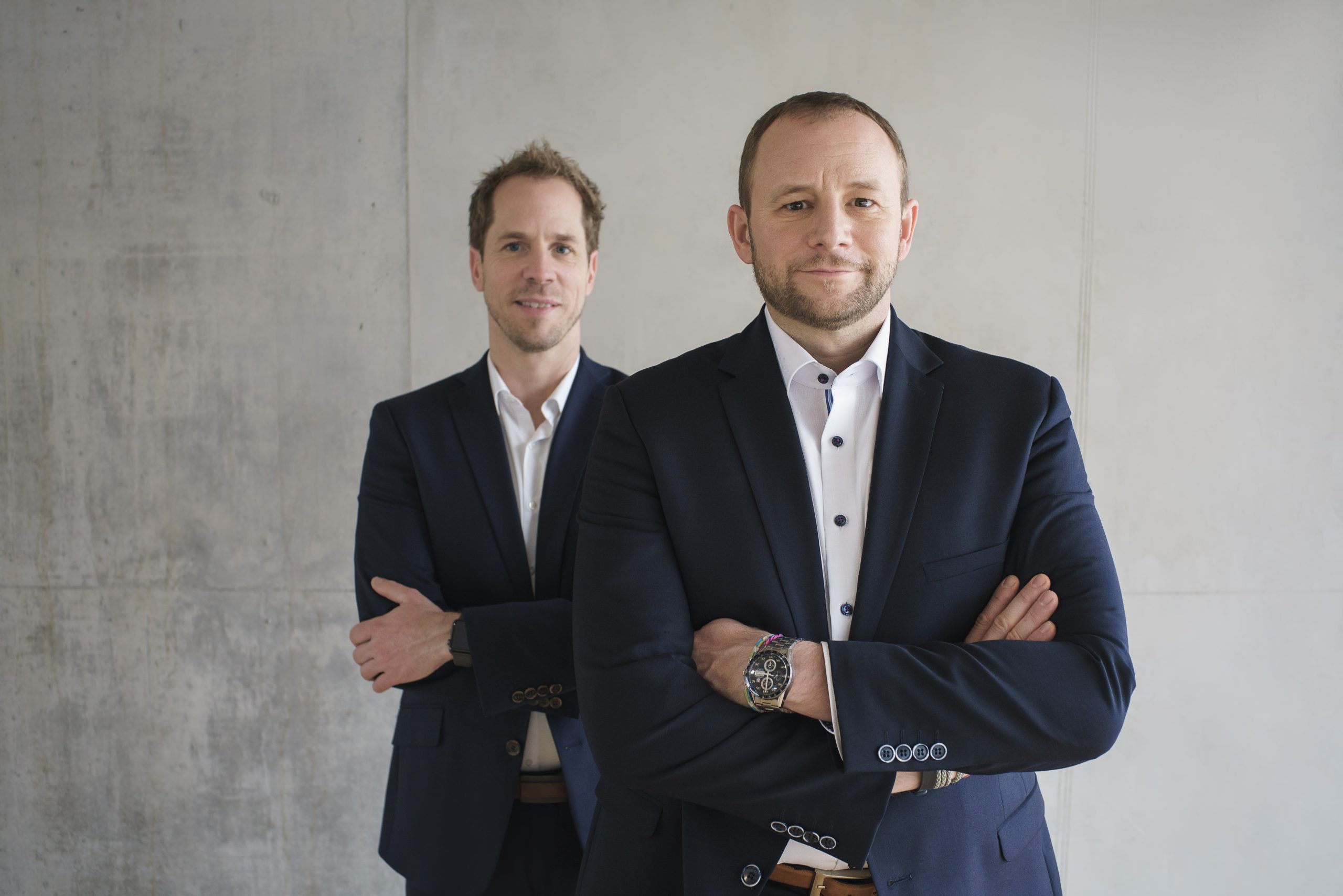 The two managing directors of virtual7 in suits, in front of a neutral background, with their arms crossed.