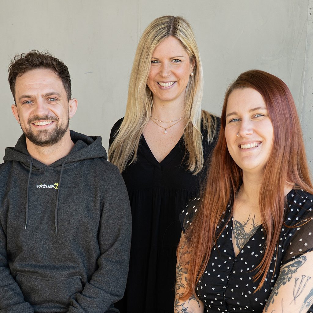 Drei Mitarbeiter des HR-Recruiting-Teams von virtual7 lächeln in die Kamera vor neutralem Hintergrund.
